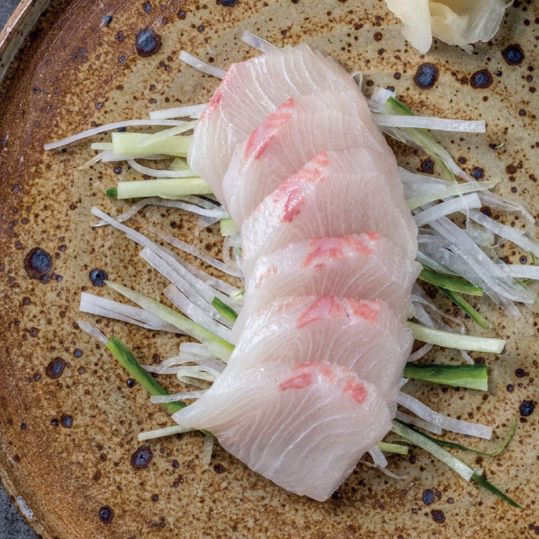 Five slices of Hiramasa (yellowtail kingfish) sashimi, bias-cut to reveal delicate pink marbling, fanned over a bed of julienned daikon and cucumber, served on a rustic speckled ceramic plate with pickled ginger in the background.