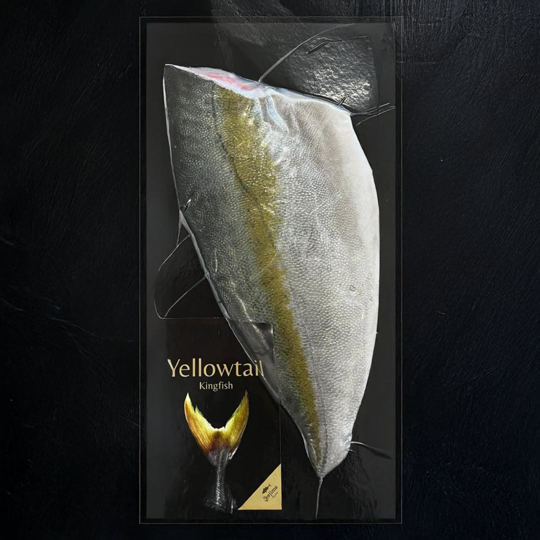 A whole Yellowtail Kingfish (Hiramasa) vacuum-sealed in premium black retail packaging, showing its distinctive silver-white belly and olive-gold lateral stripe, with the brand's signature golden tail fin logo and "Yellowtail Kingfish" label visible at the bottom.